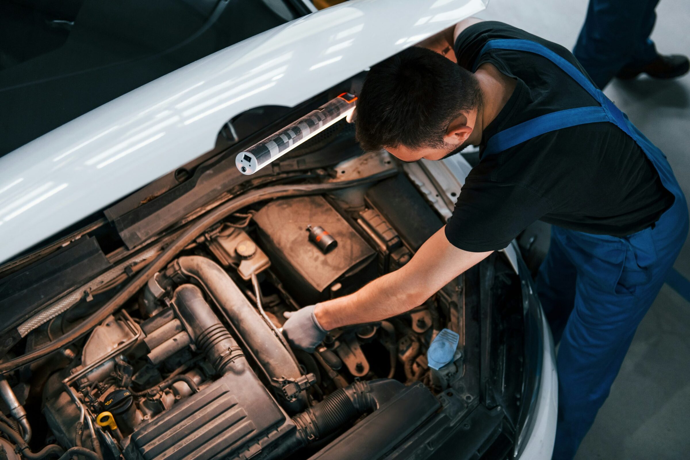 Mechanic in blue overalls checking the engine bay of a white car under a work light clipped to the open hood