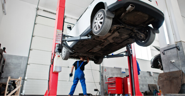 Mechanic in blue overalls working beneath a white car raised on a red two-post lift in a repair shop