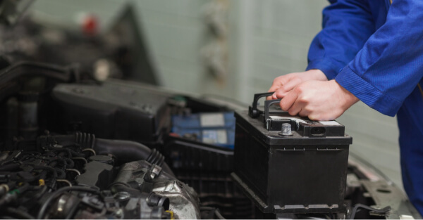 Technician in a blue uniform lifting a car battery out of an engine bay