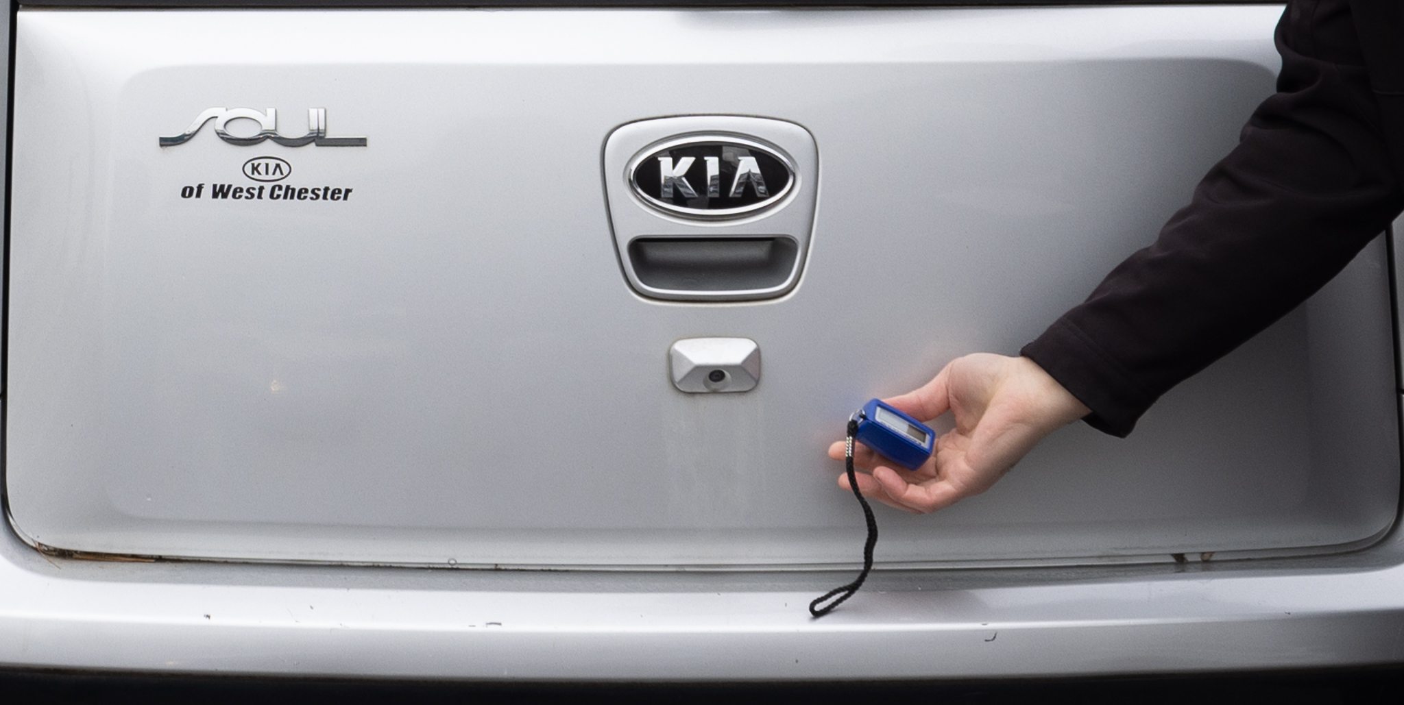 Hand holding a paint thickness gauge against the rear hatch of a silver Kia Soul