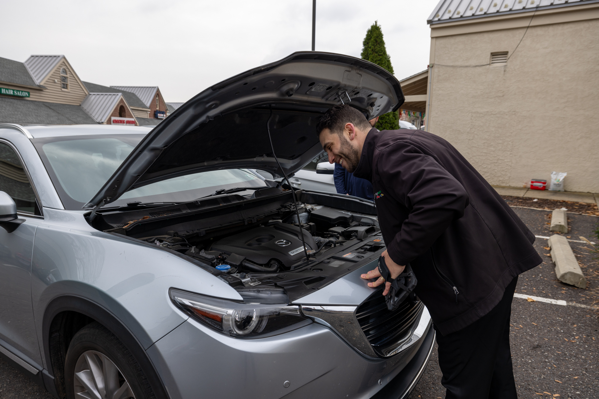 Man leaning in to look at the engine of a silver Mazda SUV with its hood open in a parking lot